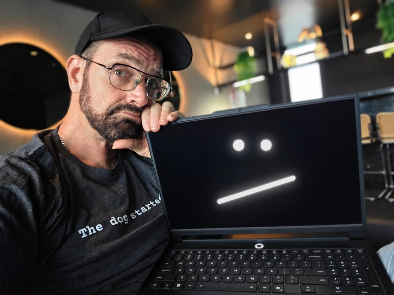 Adam in a black cap and a shirt that reads The dog started it., leaning on a laptop whose screen shows two glowing eyes and a straight mouth.