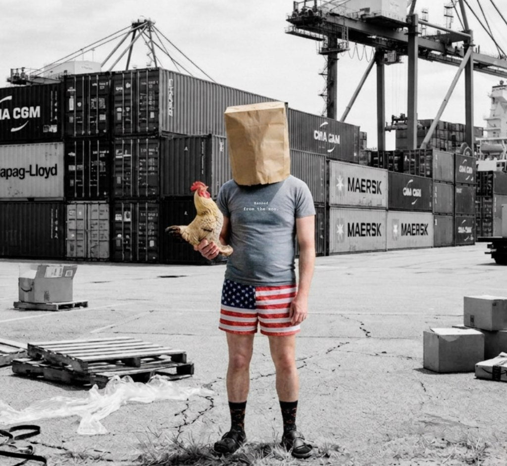 A man stands in a shipping container yard with a paper bag over his head, holding a rubber chicken. His shirt reads Banned from the zoo.